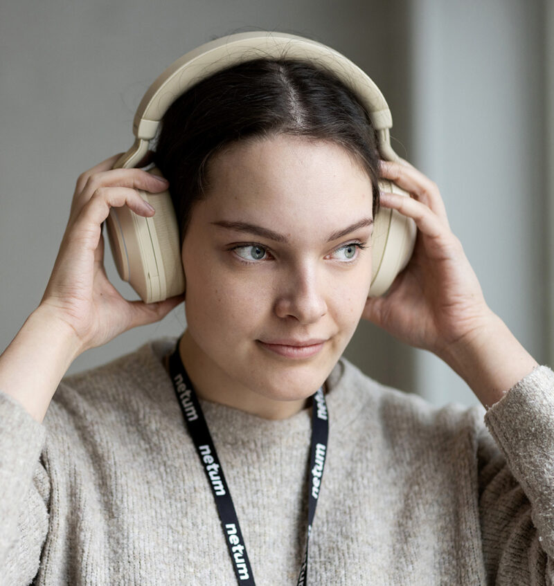 Henkilö laittaa päähänsä vaaleat over-ear-kuulokkeet, katsoo sivulle ja hymyilee kevyesti, yllään neule ja kaulassa työpaikan kaulanauha.