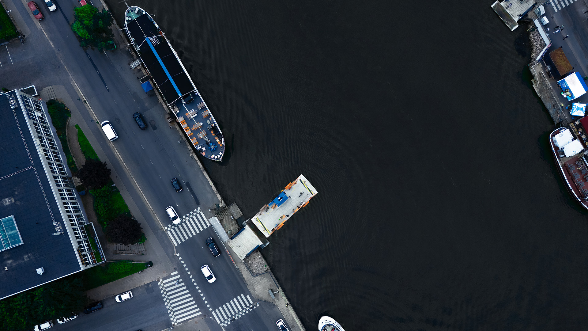 Ilmakuva joenrannasta: pieni lautta (föri) kiinnittyneenä laiturissa tumman veden reunassa, viereisellä satamalaiturilla suurempi alus ja rantakadulla suojatie, autoja ja rakennuksia.