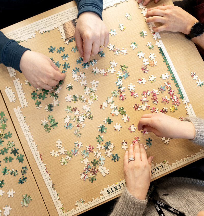 Yläkuvasta näkyvä pöytä, jossa neljä ihmistä kokoavat palapeliä; palapelin reunat osittain koottu ja keskellä levällään värikkäitä paloja, näkyvissä vain kädet ja erilaiset hihat.