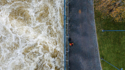 Ilmakuva koskesta ja rinnakkaisesta kävelypolusta: vasemmalla kuohuva virta, keskellä polun reunalla kaide ja yksi kävelijä oranssissa takissa, oikealla nurmikkoa, puita ja katuvaloja