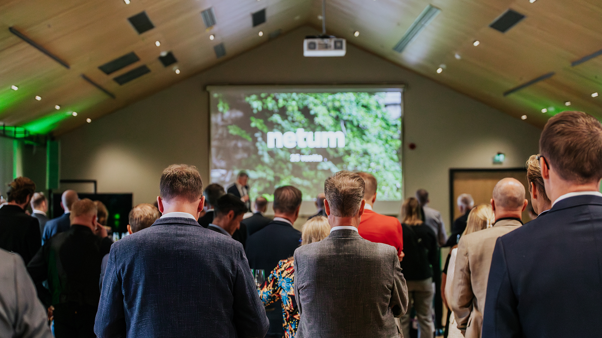 Ihmisiä seisomassa etualalla selin kameralle katsomassa esiintymislavaa ja suurta valkokangasta, jolla näkyy "netum"-logo vihreällä kasvikuvataustalla yritystapahtumassa kirkkaasti valaistussa salissa.