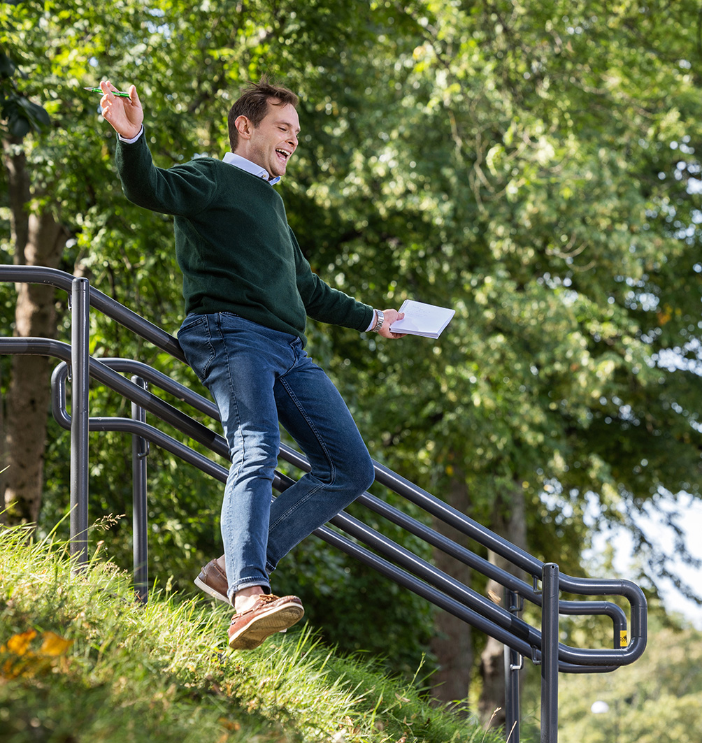 Hymyilevä henkilö laskeutuu pientä nurmista rinnettä ja kaidetta pitkin pitäen valkoista paperia, taustalla vehreää puistomaisemaa.