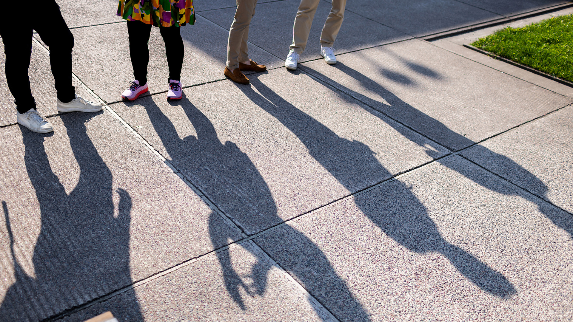 Ihmisten jalat ja pitkät varjot auringossa laatoitetulla kävelytielellä; näkyvissä eriväriset kengät, osa värikkäästä hameesta ja nurmikon reuna.