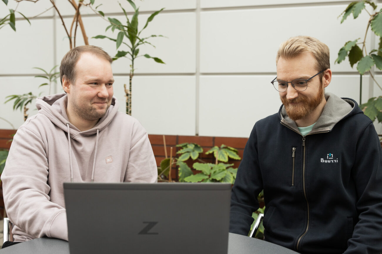 Two smiling web developers sitting on the front of a laptop. Plants on the background.