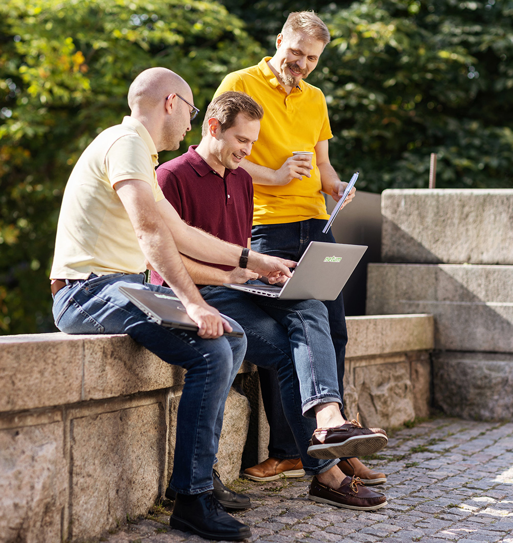 Kolme aikuista miestä rentoissa vaatteissa istuu ja seisoo kivisellä reunalla puiston reunassa; keskellä istuva mies käyttää kannettavaa tietokonetta, vieressä istuva mies katsoo näyttöä ja takana seisova mies pitelee kahvimukia ja vihkoa hymyillen.