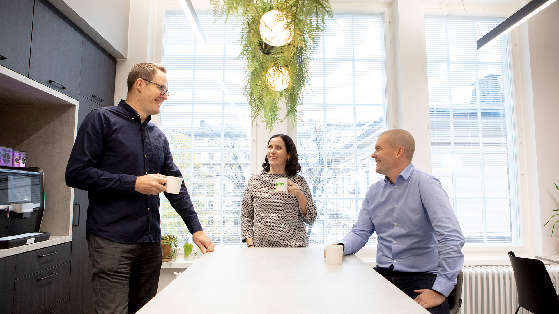 Kolme toimistotyöntekijää seisoo keittiösaarekkeella toimiston taukotilassa, pitävät kahvimukeja ja keskustelevat hymyillen suurten ikkunoiden ja riippuvan viherkasvin edustalla.