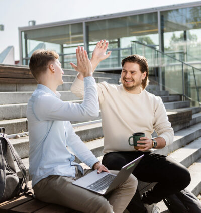 Kaksi opiskelijaa istuu ulkona portailla, toisella kannettava tietokone sylissä ja toisella kahvimuki, he hymyilevät ja antavat ylävitosen kampuksen portailla modernin lasirakennuksen edessä.
