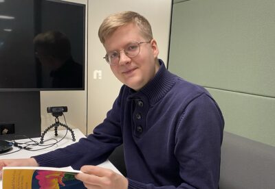 Iloinen sulautettujen järjestelmien kehittäjä katsoo kameraan ja selaa samalla kirjaa.